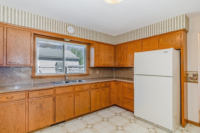 kitchen featuring freestanding refrigerator, light floors, wallpapered walls, brown cabinets, and a sink