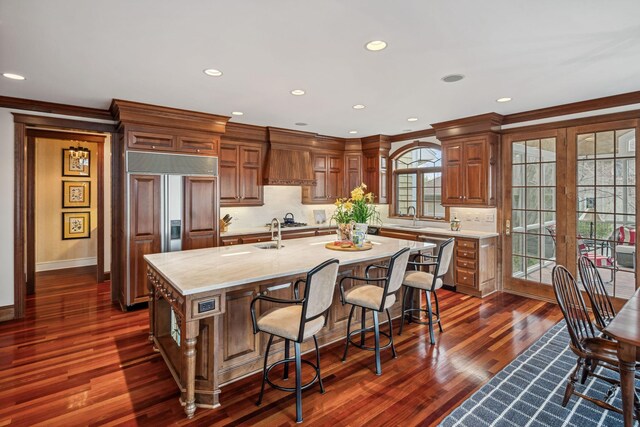 kitchen with decorative backsplash, paneled refrigerator, a sink, premium range hood, and an island with sink