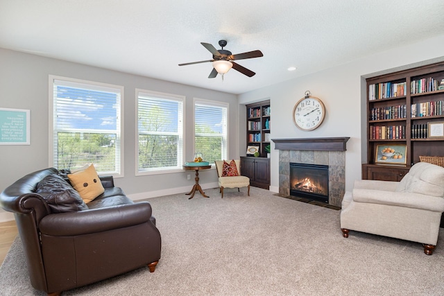 carpeted living area featuring a tiled fireplace, built in shelves, a ceiling fan, and baseboards