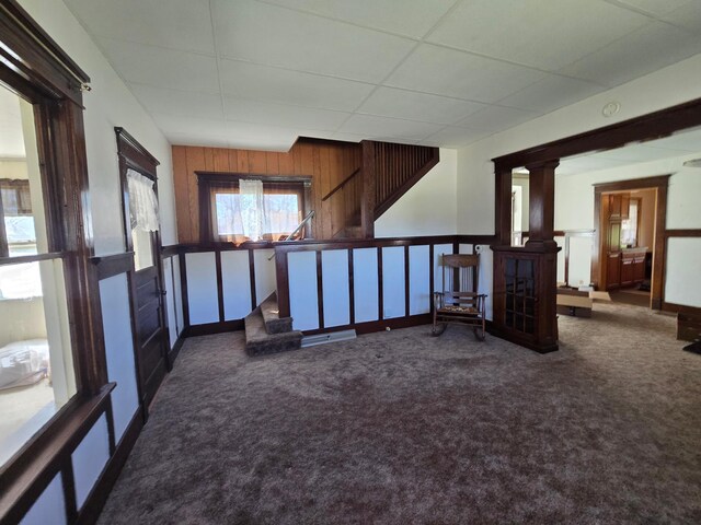carpeted empty room with wood walls, a drop ceiling, and stairway
