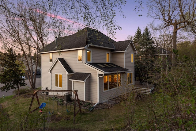 back of house at dusk with central AC unit, roof with shingles, and a yard