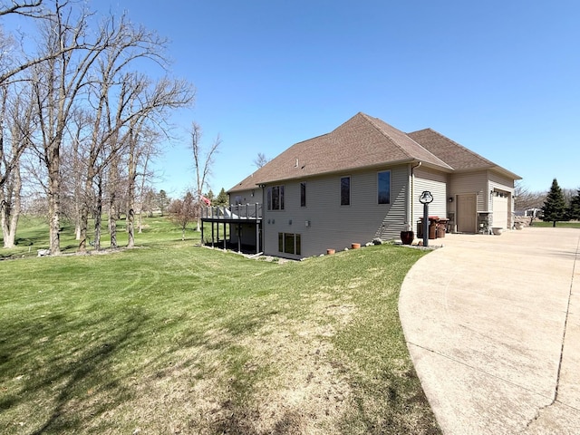 view of side of home with an attached garage, a shingled roof, a deck, concrete driveway, and a lawn