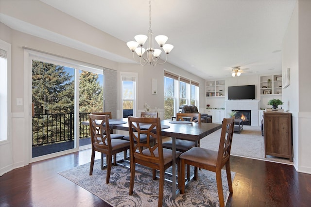 dining area with a glass covered fireplace, ceiling fan with notable chandelier, dark wood-style floors, and built in shelves