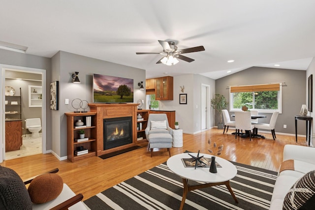 living area with ceiling fan, a glass covered fireplace, baseboards, light wood-type flooring, and vaulted ceiling