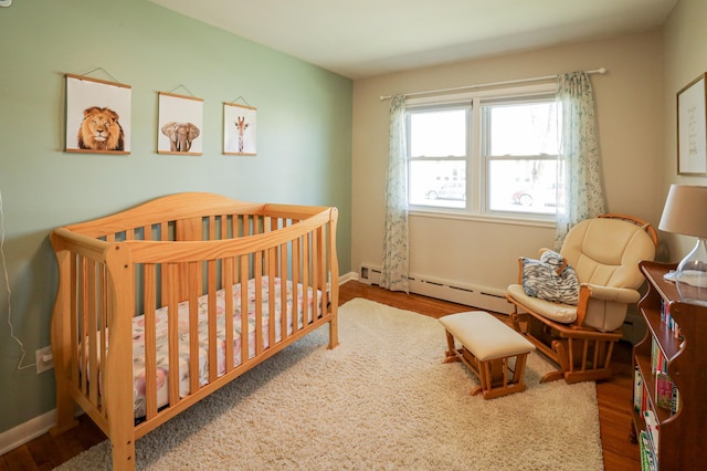 bedroom featuring a nursery area, baseboards, a baseboard heating unit, and wood finished floors
