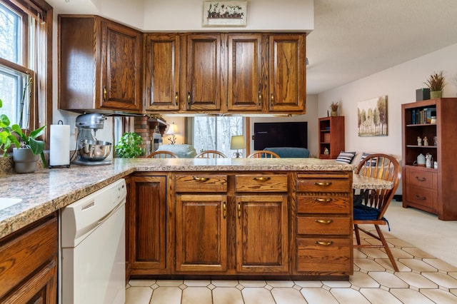 kitchen with white dishwasher, light colored carpet, light stone counters, brown cabinetry, and a peninsula