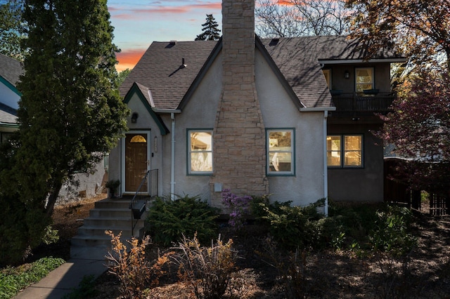 tudor home featuring stucco siding, a balcony, a shingled roof, and a chimney