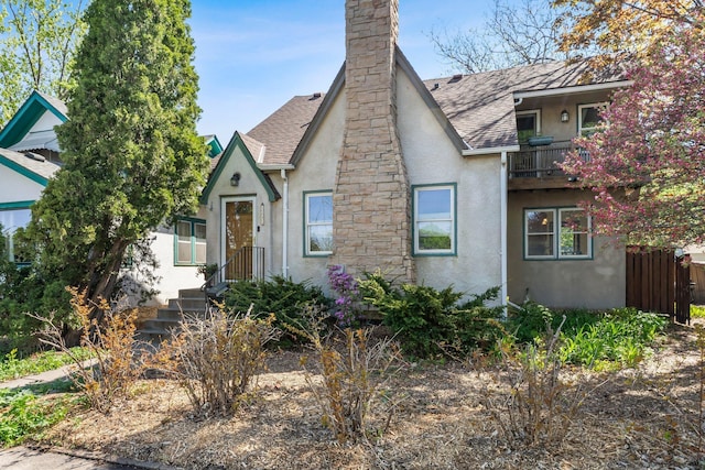 tudor house with stucco siding, a shingled roof, a balcony, fence, and a chimney
