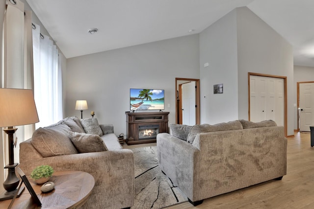 living area featuring light wood-type flooring, a glass covered fireplace, and high vaulted ceiling