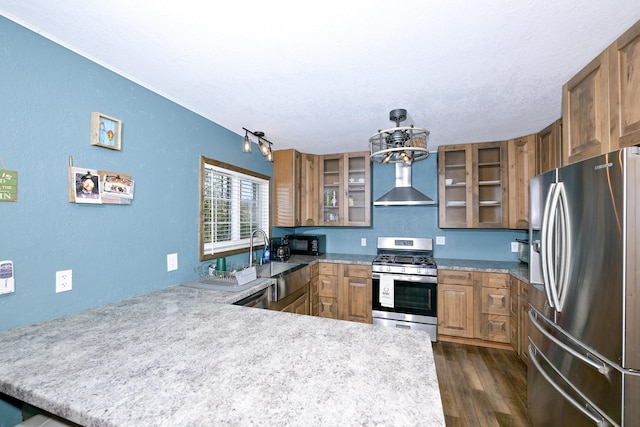 kitchen with a peninsula, a chandelier, wall chimney exhaust hood, brown cabinets, and stainless steel appliances