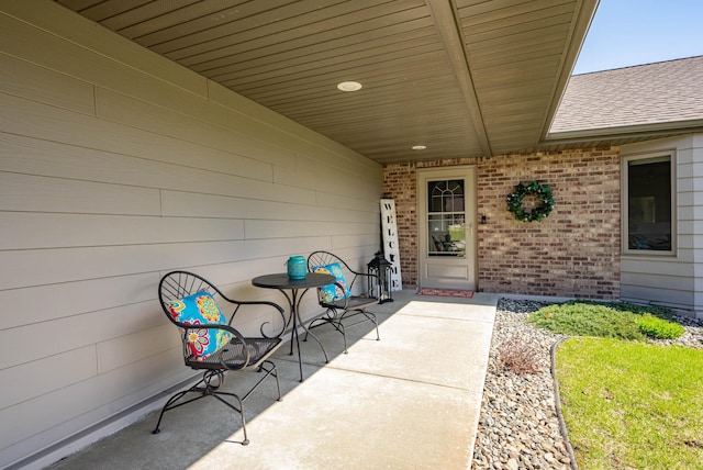 view of exterior entry featuring brick siding, a shingled roof, and a patio area