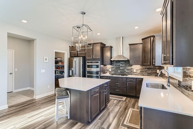 kitchen featuring wall chimney range hood, decorative backsplash, dark brown cabinetry, appliances with stainless steel finishes, and a sink