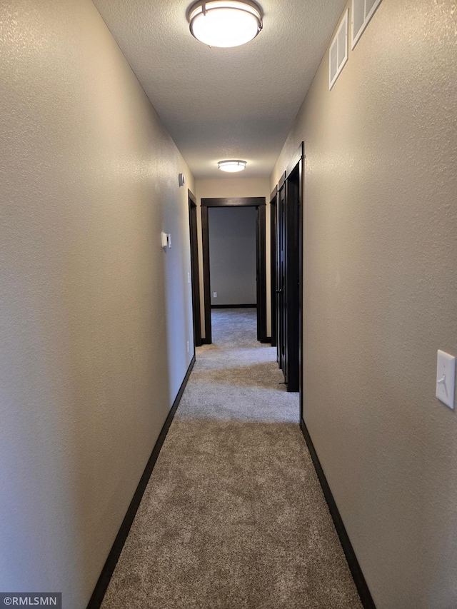 hallway with a textured ceiling, visible vents, light colored carpet, baseboards, and a textured wall