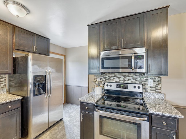 kitchen featuring stainless steel appliances, wainscoting, dark brown cabinets, light stone counters, and decorative backsplash