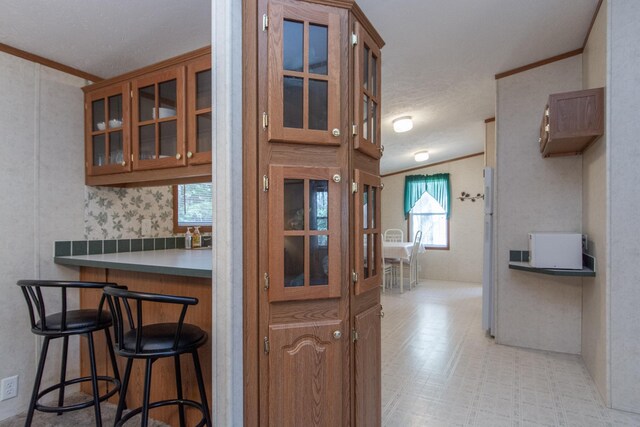 kitchen with brown cabinetry, ornamental molding, glass insert cabinets, light floors, and a kitchen bar