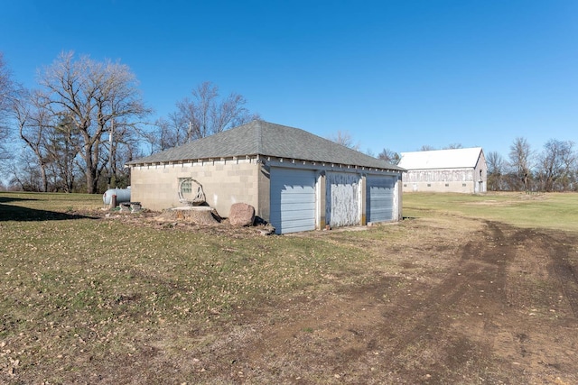 view of side of home with roof with shingles, a garage, concrete block siding, an outbuilding, and a yard