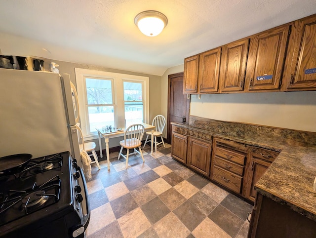 kitchen featuring black range with gas cooktop, brown cabinetry, stone finish flooring, dark countertops, and freestanding refrigerator