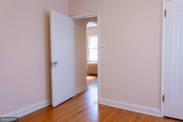 unfurnished room featuring radiator heating unit, arched walkways, light wood-style flooring, and baseboards