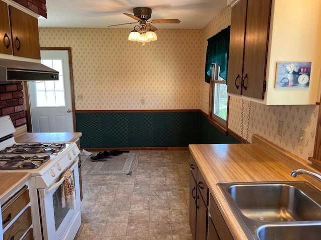 kitchen with white gas range oven, a sink, light countertops, wallpapered walls, and under cabinet range hood