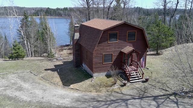 view of front of house with a gambrel roof, roof with shingles, a chimney, and a forest view