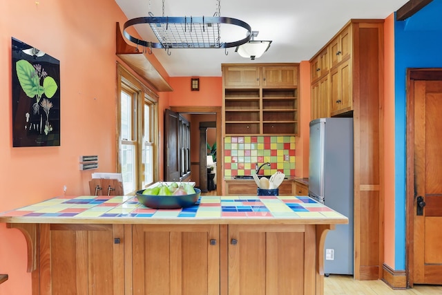 kitchen with decorative backsplash, tile counters, freestanding refrigerator, and brown cabinetry
