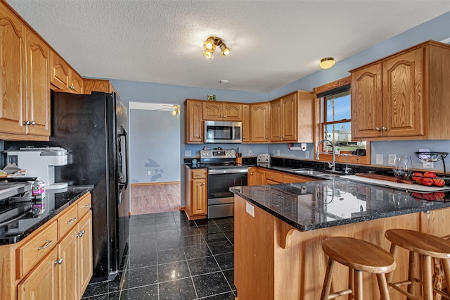 kitchen with a sink, dark stone countertops, stainless steel appliances, a peninsula, and a kitchen bar