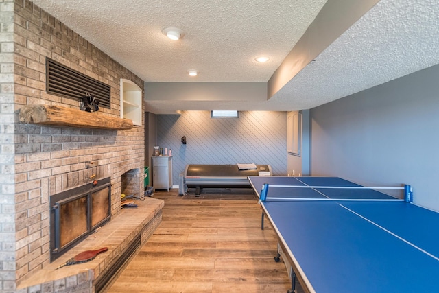 rec room featuring light wood-style flooring, a textured ceiling, and a brick fireplace