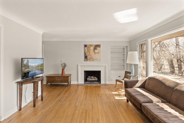living room featuring a brick fireplace, baseboards, built in shelves, and light wood-type flooring
