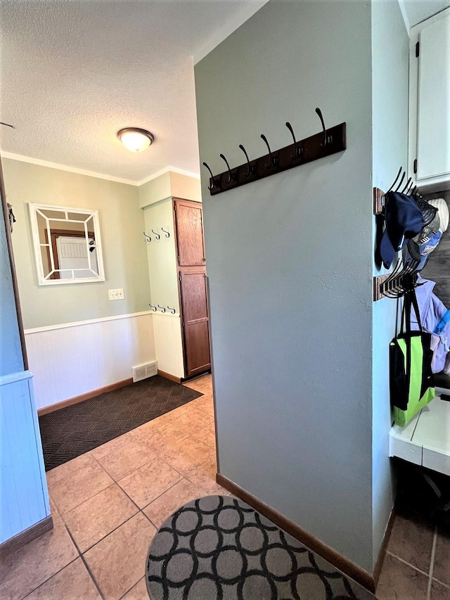 hall featuring wainscoting, light tile patterned flooring, a textured ceiling, ornamental molding, and baseboards