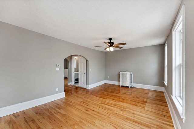 spare room featuring ceiling fan, arched walkways, radiator heating unit, baseboards, and light wood-type flooring