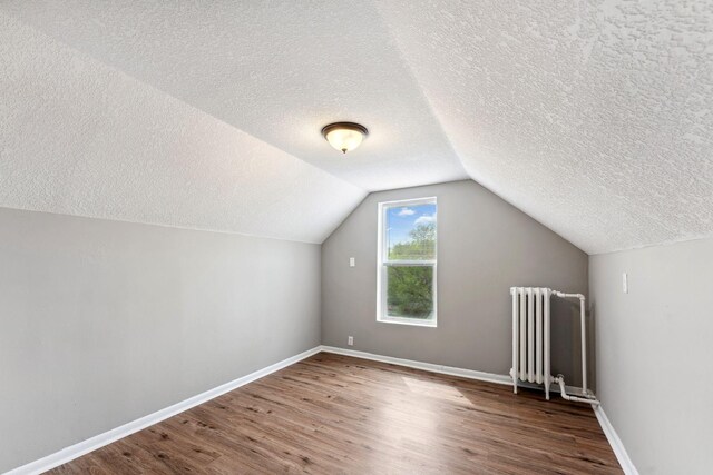 additional living space with radiator, wood finished floors, baseboards, a textured ceiling, and vaulted ceiling
