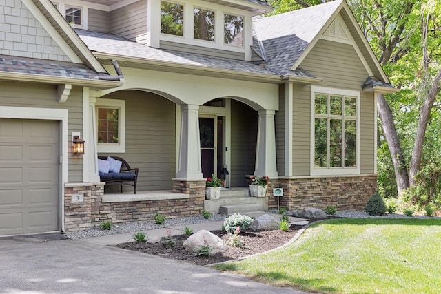 entrance to property with covered porch, roof with shingles, stone siding, an attached garage, and a yard