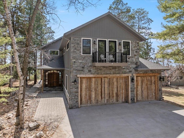 view of front of house with stone siding, an attached garage, a balcony, a shingled roof, and driveway