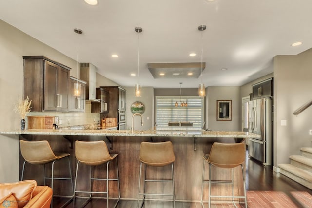 kitchen with light stone counters, a peninsula, wall chimney exhaust hood, appliances with stainless steel finishes, and dark wood-style floors