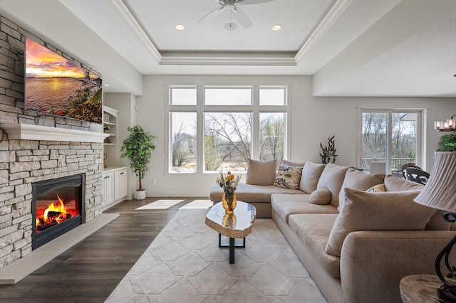 living room featuring ceiling fan, a stone fireplace, wood finished floors, a tray ceiling, and recessed lighting