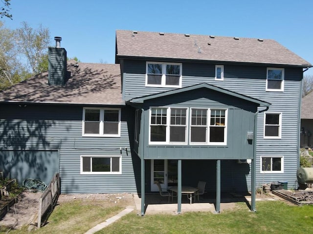 back of house featuring a patio, a chimney, heating fuel, a shingled roof, and a yard