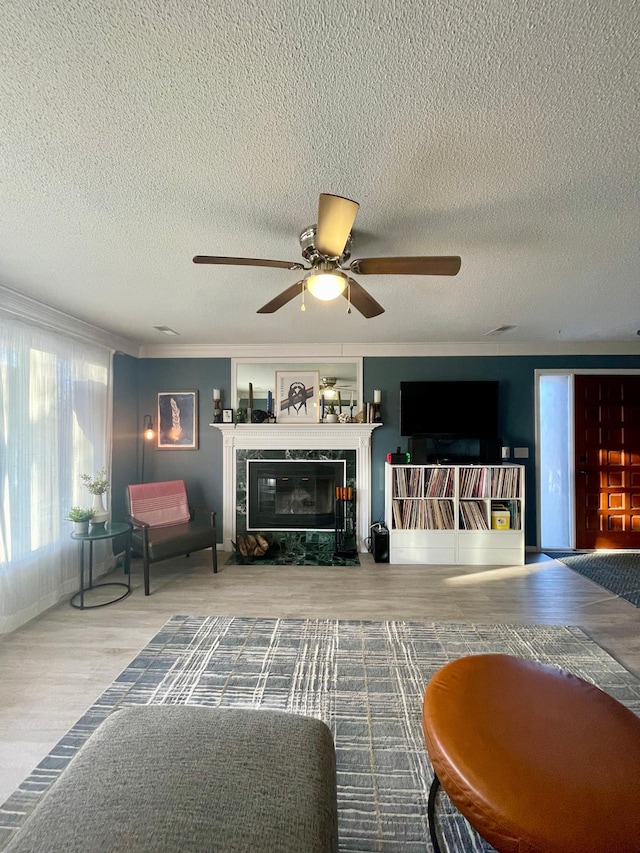 living room featuring ceiling fan, a textured ceiling, a tiled fireplace, wood finished floors, and ornamental molding