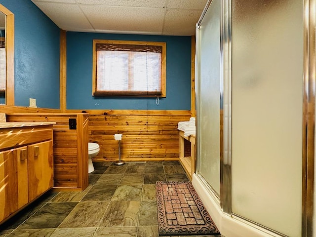 full bathroom featuring wooden walls, toilet, stone finish flooring, a drop ceiling, and a stall shower