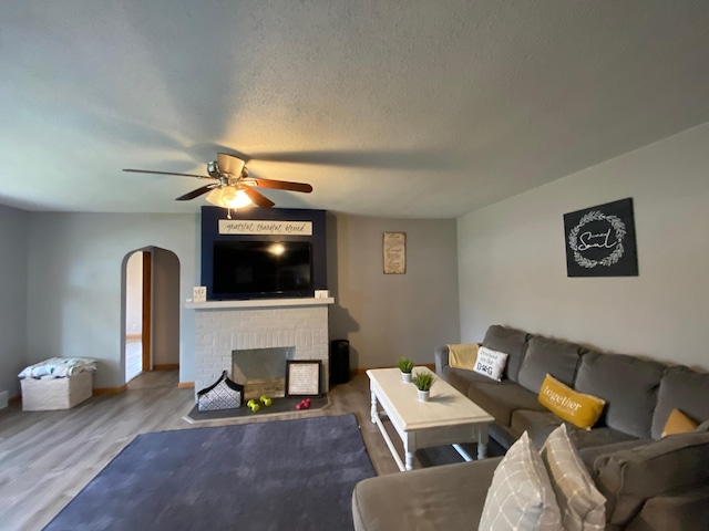 living area featuring ceiling fan, a textured ceiling, arched walkways, a brick fireplace, and wood finished floors
