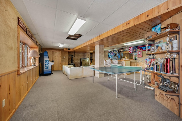 rec room with carpet flooring, a drop ceiling, wood walls, and a wood stove