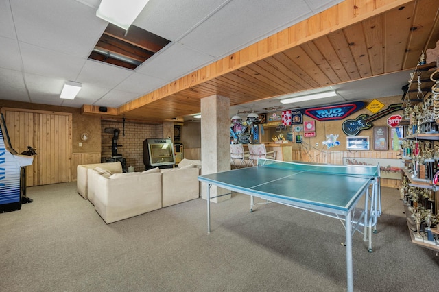 rec room with a wood stove, a paneled ceiling, carpet flooring, and wood walls