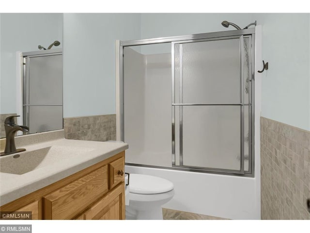 bathroom featuring shower / bath combination with glass door, vanity, toilet, a wainscoted wall, and tile walls