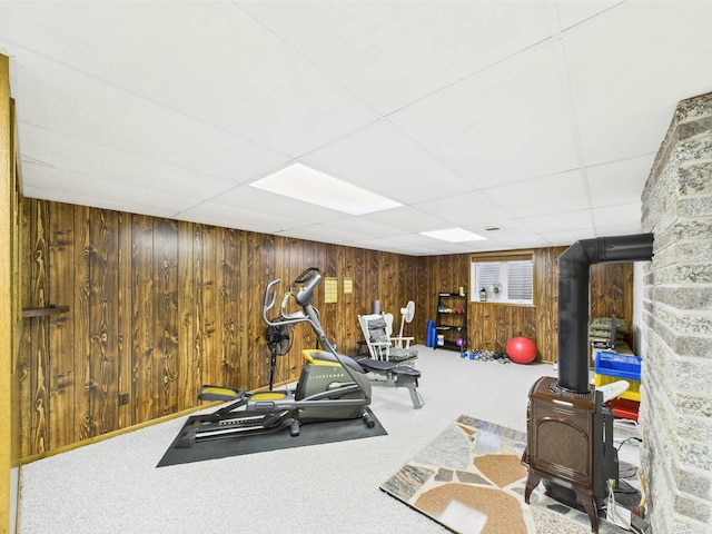 workout area with a wood stove, carpet, a drop ceiling, and wood walls
