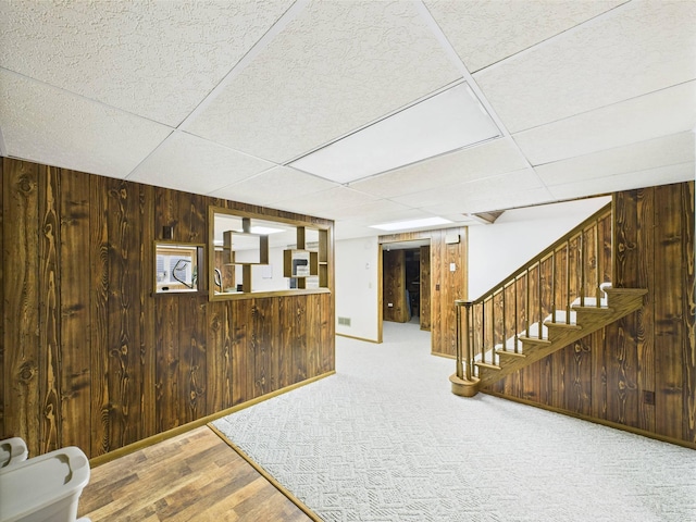 finished basement featuring visible vents, a paneled ceiling, wood walls, baseboards, and stairs
