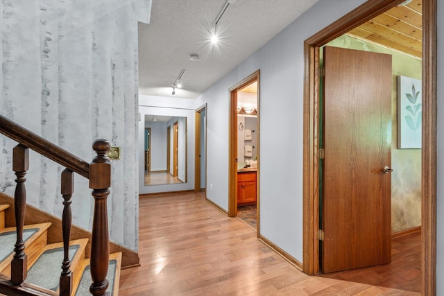 hallway featuring rail lighting, light wood-type flooring, stairway, and baseboards