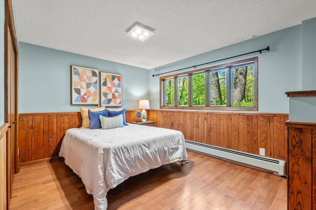 bedroom featuring a baseboard heating unit, a wainscoted wall, a textured ceiling, wooden walls, and wood finished floors