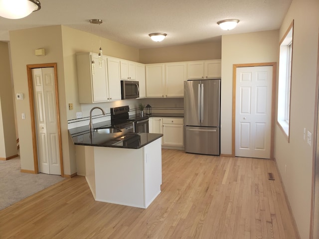 kitchen featuring stainless steel appliances, a peninsula, dark countertops, light wood finished floors, and a sink