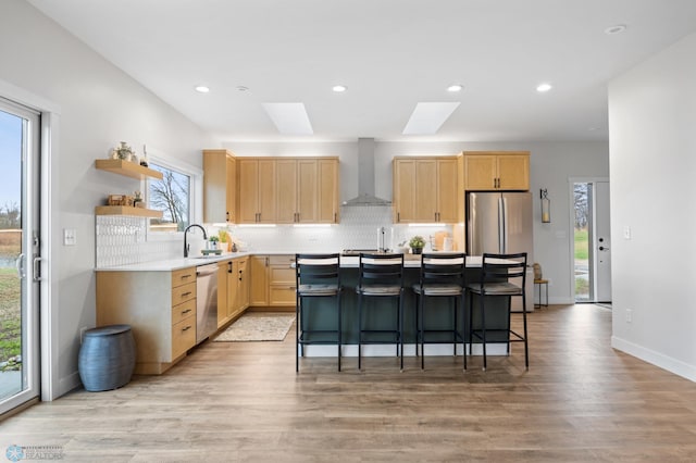 kitchen with light brown cabinets, a center island, wall chimney exhaust hood, a skylight, and stainless steel appliances