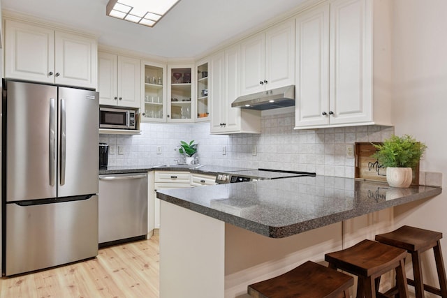 kitchen with a peninsula, glass insert cabinets, under cabinet range hood, light wood finished floors, and stainless steel appliances