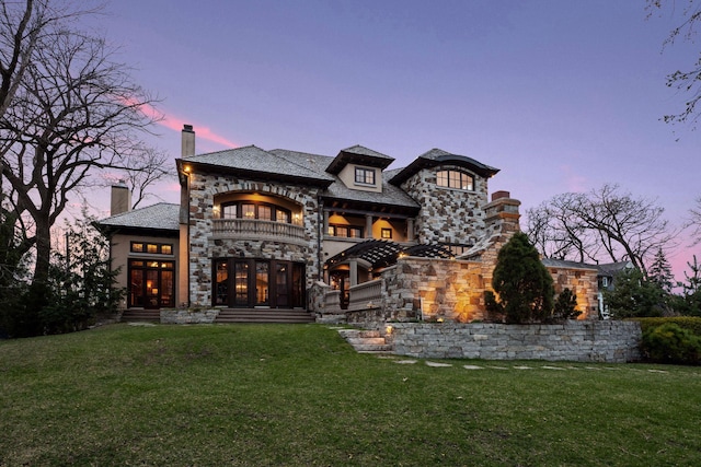back of house at dusk with a lawn, a balcony, and stone siding